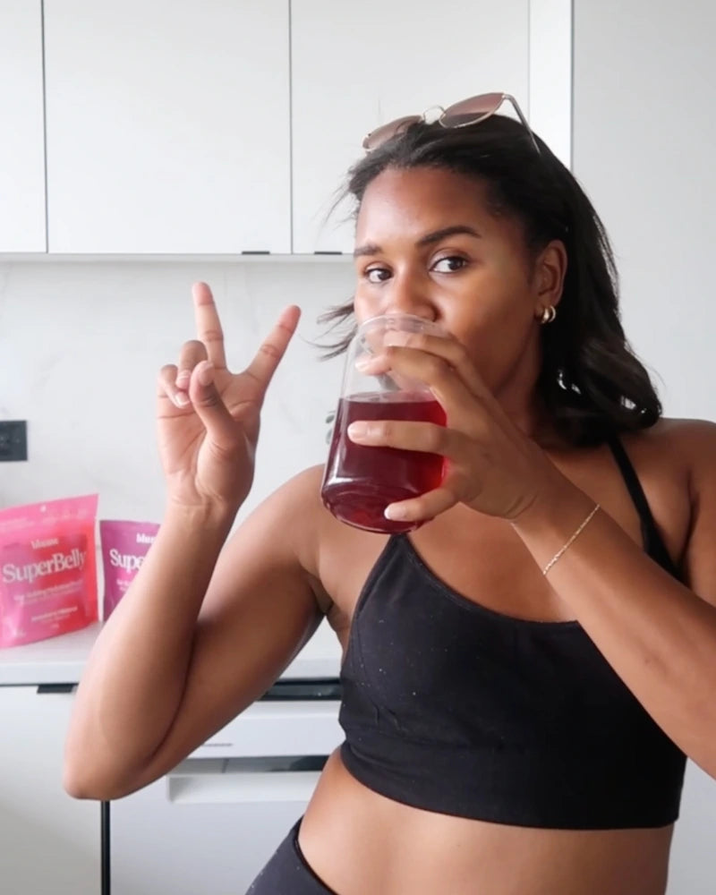A person in a black tank top drinks a red beverage from a glass, flashing a peace sign. In the background, two pink pouches labeled SuperBelly are on a kitchen counter.